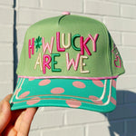 Green cap with colorful text 'HOW LUCKY ARE WE' held against a white brick wall.