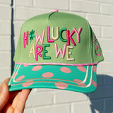 Green cap with colorful text 'HOW LUCKY ARE WE' held against a white brick wall.