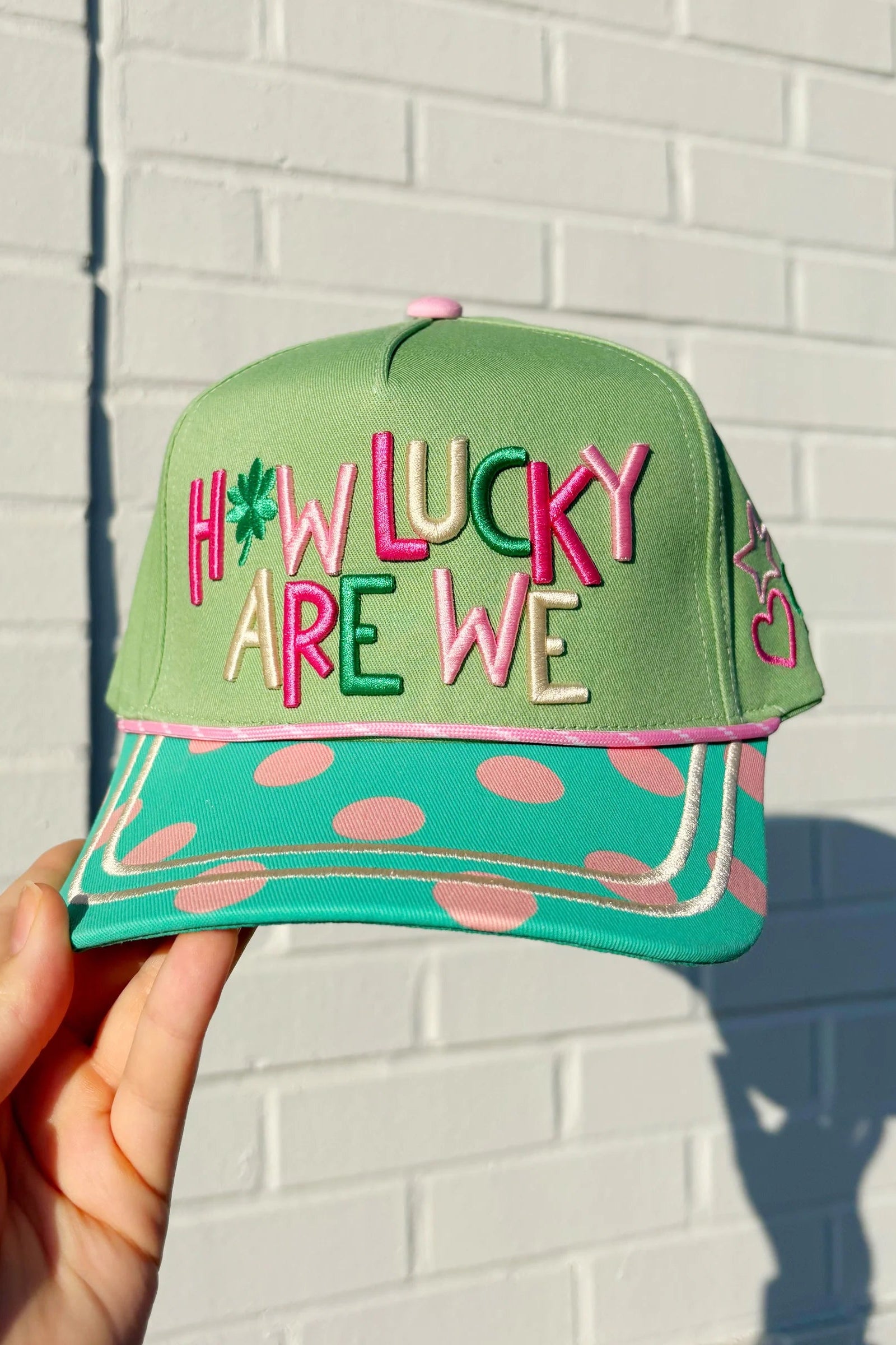 Green cap with colorful text 'HOW LUCKY ARE WE' held against a white brick wall.