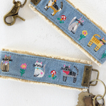 Denim key fob with embroidered designs of cats and flowers on a white background