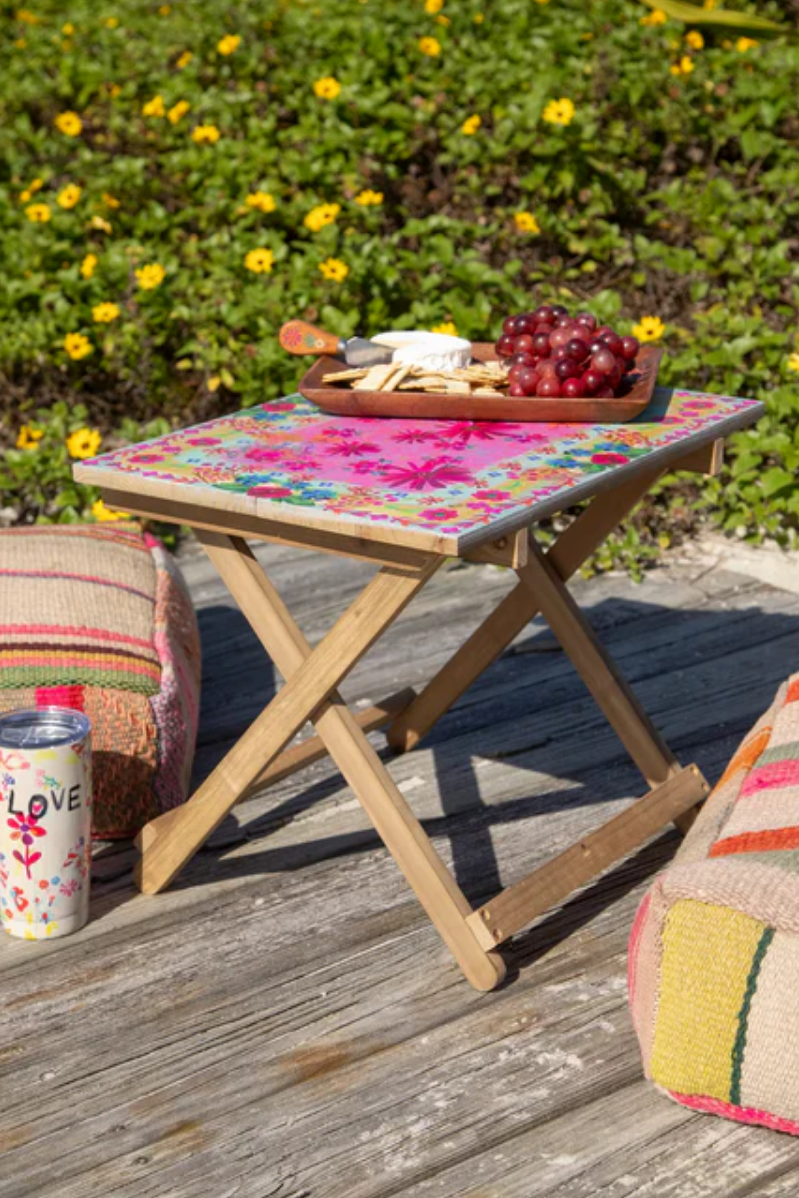 Small folding table with a floral cover outdoors, surrounded by greenery and a can.
