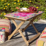 Small folding table with a floral cover outdoors, surrounded by greenery and a can.