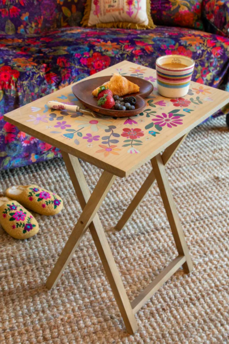 Small wooden table with floral design on a living room floor, set with a plate of food and cups.