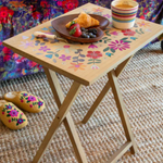 Small wooden table with floral design on a living room floor, set with a plate of food and cups.