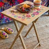 Small wooden table with floral design on a living room floor, set with a plate of food and cups.