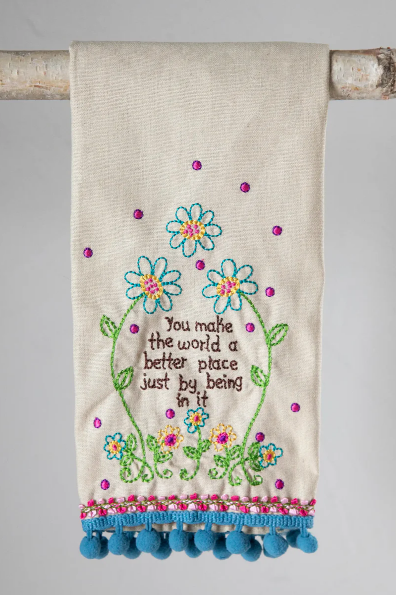 Decorative towel with floral embroidery and pom-pom trim on a wooden stick against a gray background