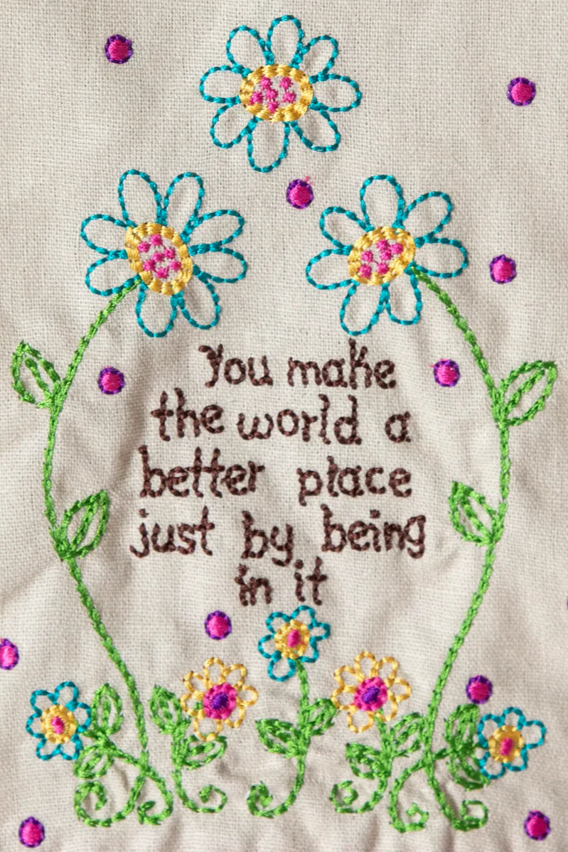Embroidered text with flowers on a beige background reading, "You make the world a better place just by being in it"