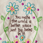 Embroidered text with flowers on a beige background reading, "You make the world a better place just by being in it"