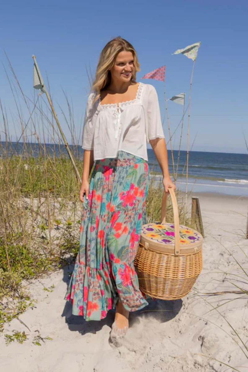 Woman on a beach holding a picnic basket, wearing a floral skirt and white top.