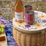 Picnic basket with a bottle of wine, cups, and snacks on a colorful tablecloth.