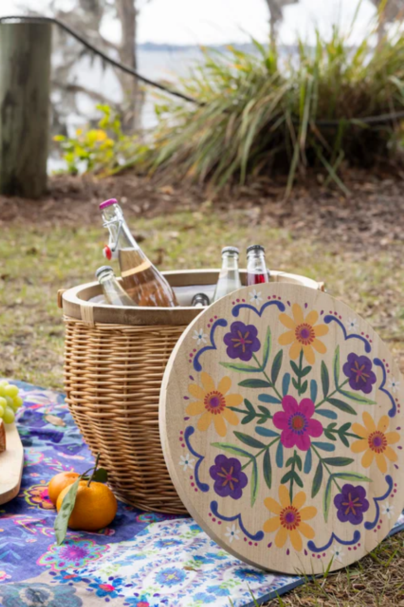 Picnic setup with a wicker basket, drinks, and a decorative plate with floral design outdoors.