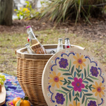 Picnic setup with a wicker basket, drinks, and a decorative plate with floral design outdoors.