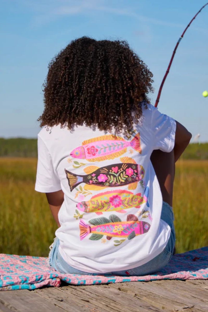Person wearing a t-shirt with colorful fish design, sitting by a body of water.