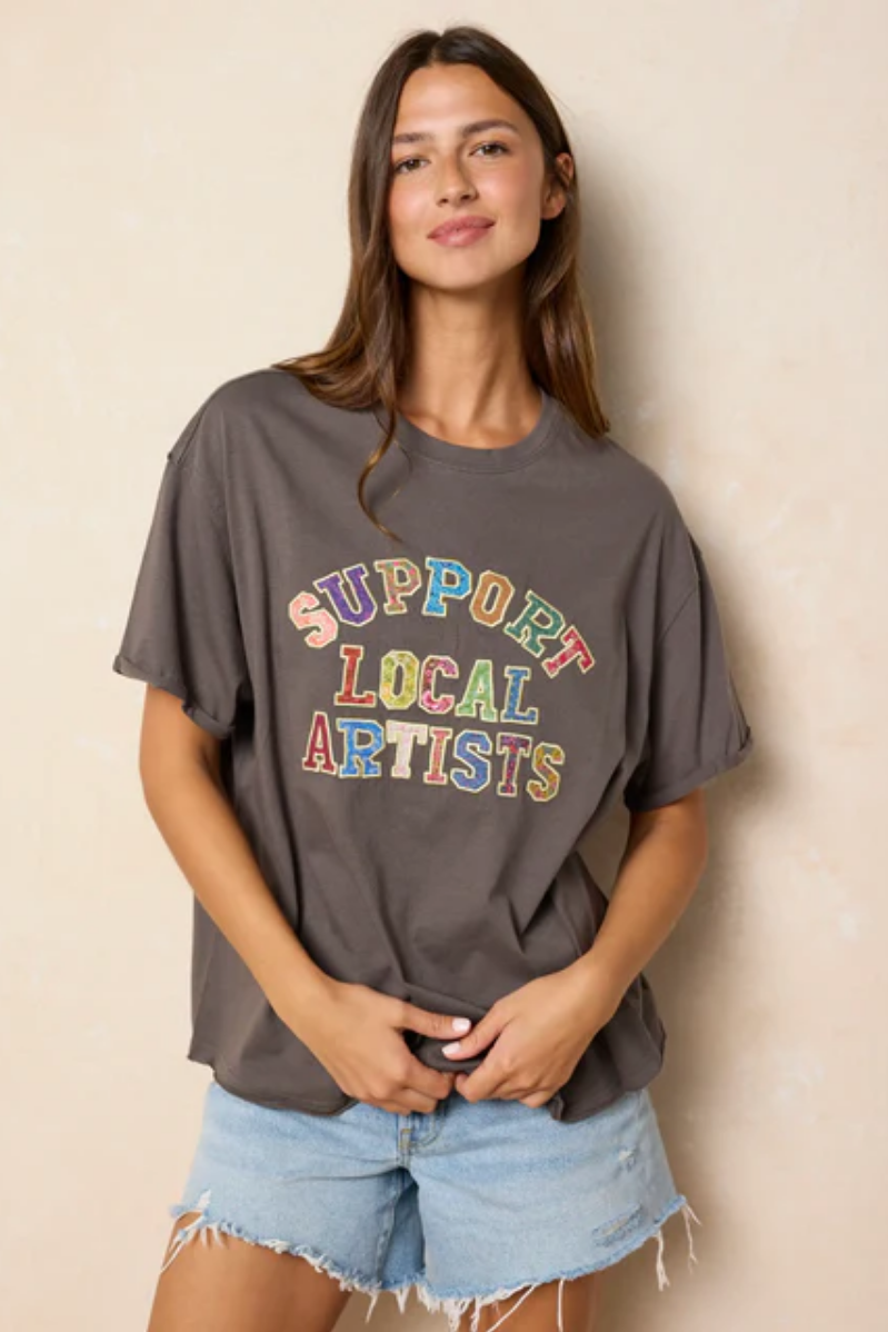 Woman wearing a gray t-shirt with 'Support Local Artists' text in colorful letters against a beige background