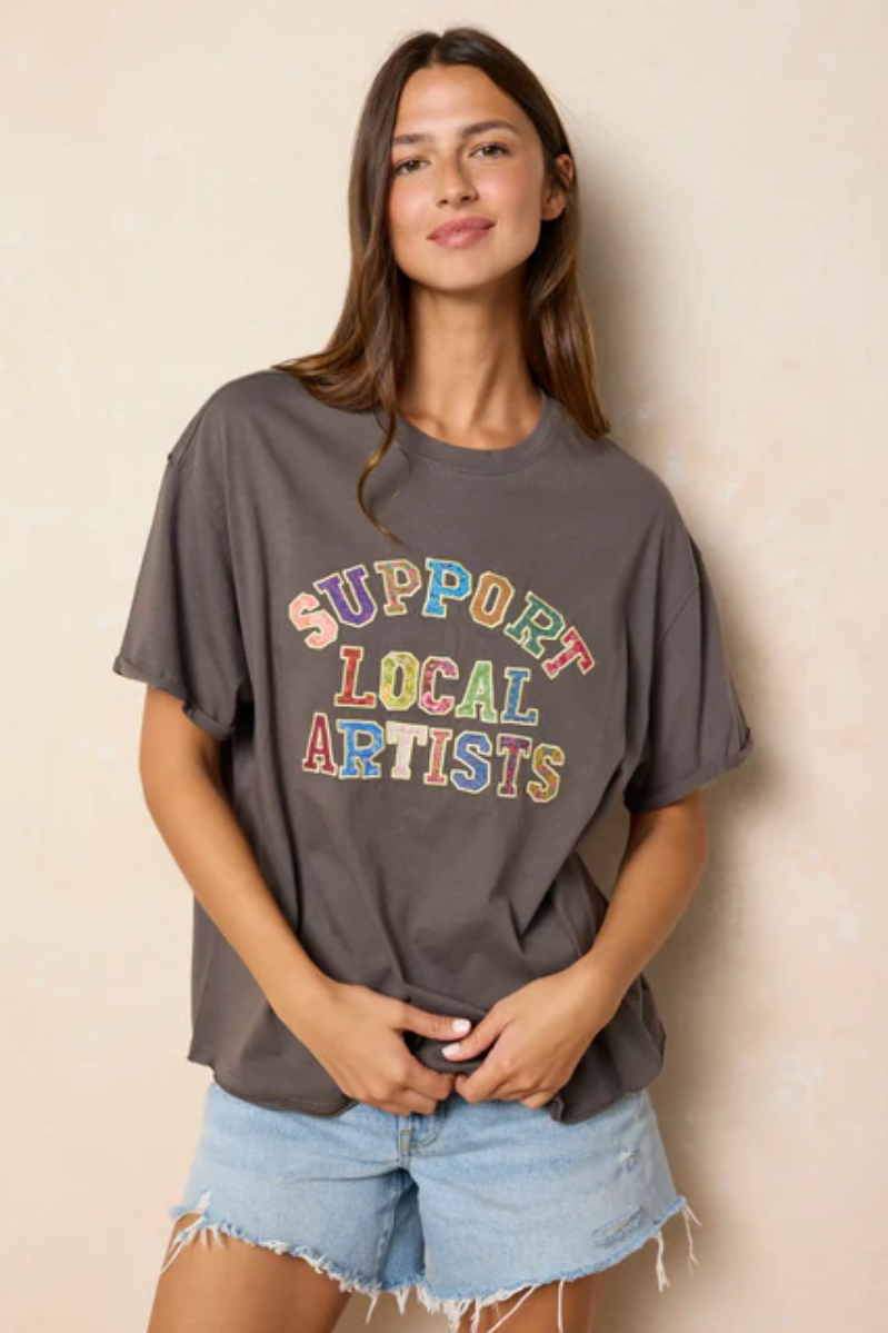 Woman wearing a gray t-shirt with 'Support Local Artists' text in colorful letters against a beige background