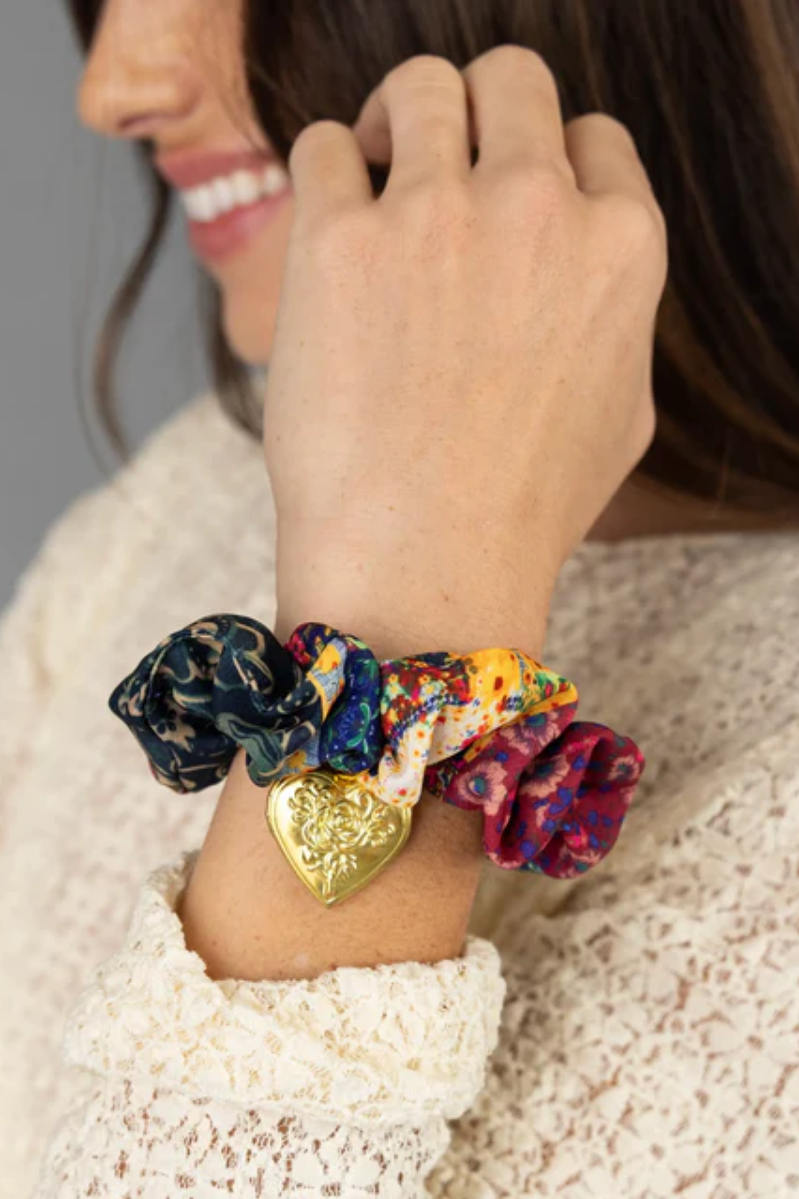 Close-up of a hand wearing colorful scrunchies with a gold heart charm on a neutral background