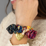 Close-up of a hand wearing colorful scrunchies with a gold heart charm on a neutral background