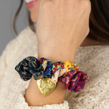 Close-up of a hand wearing colorful scrunchies with a gold heart charm on a neutral background