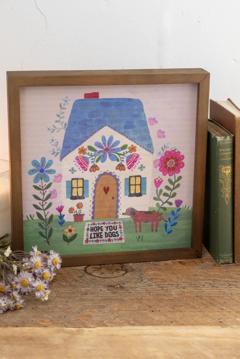 Decorative framed picture of a house with flowers and a dog on a wooden surface.