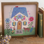 Decorative framed picture of a house with flowers and a dog on a wooden surface.