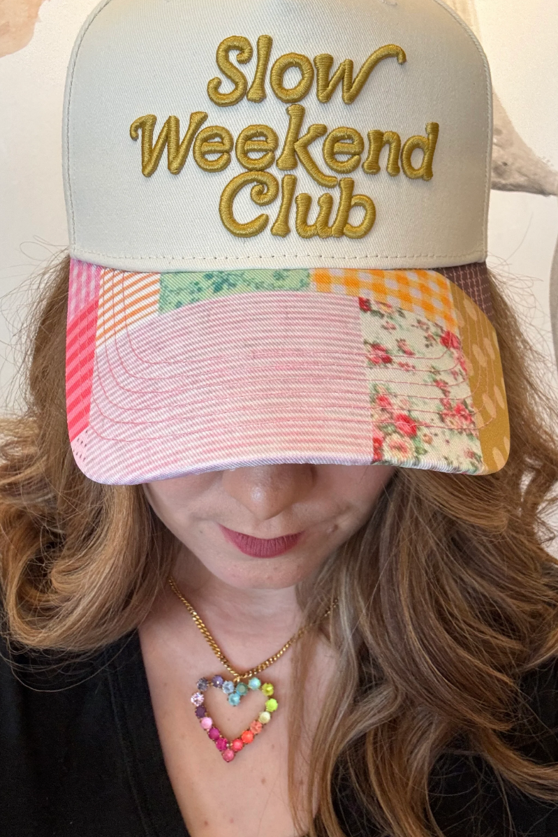 Woman wearing a cream/patchwork trucker hat that says, Slow Weekend Club.