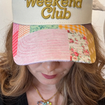 Woman wearing a cream/patchwork trucker hat that says, Slow Weekend Club.