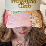 Woman wearing a cream/patchwork trucker hat that says, Slow Weekend Club.