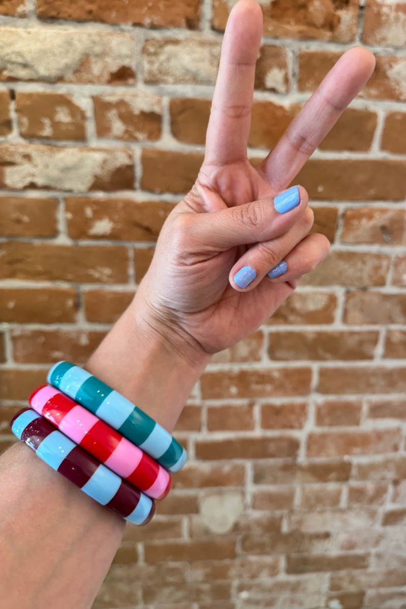 Woman wearing funky, chunky Color Crush Bangles in pink, green and brown, giving the peace sign against a brick wall. Super Groovy.