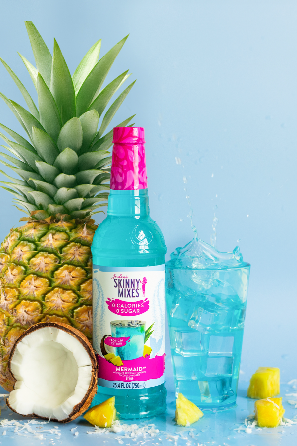 Bottle of Sunny Mixes Caipirinha Mix with a pineapple, coconut, and blue cocktail on a light blue background