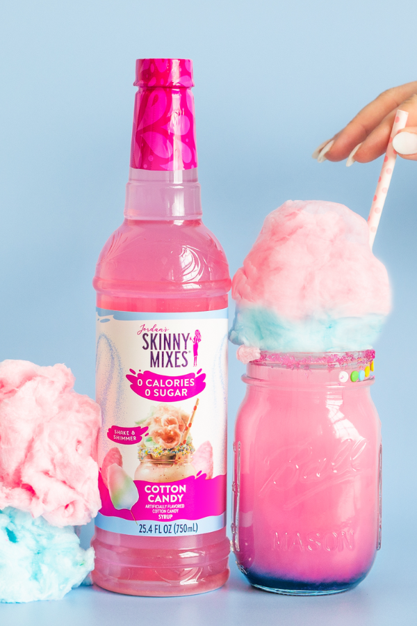 Bottle of Skinny Mixes Cotton Candy syrup with a pink cotton candy drink and cotton candy on a light blue background.