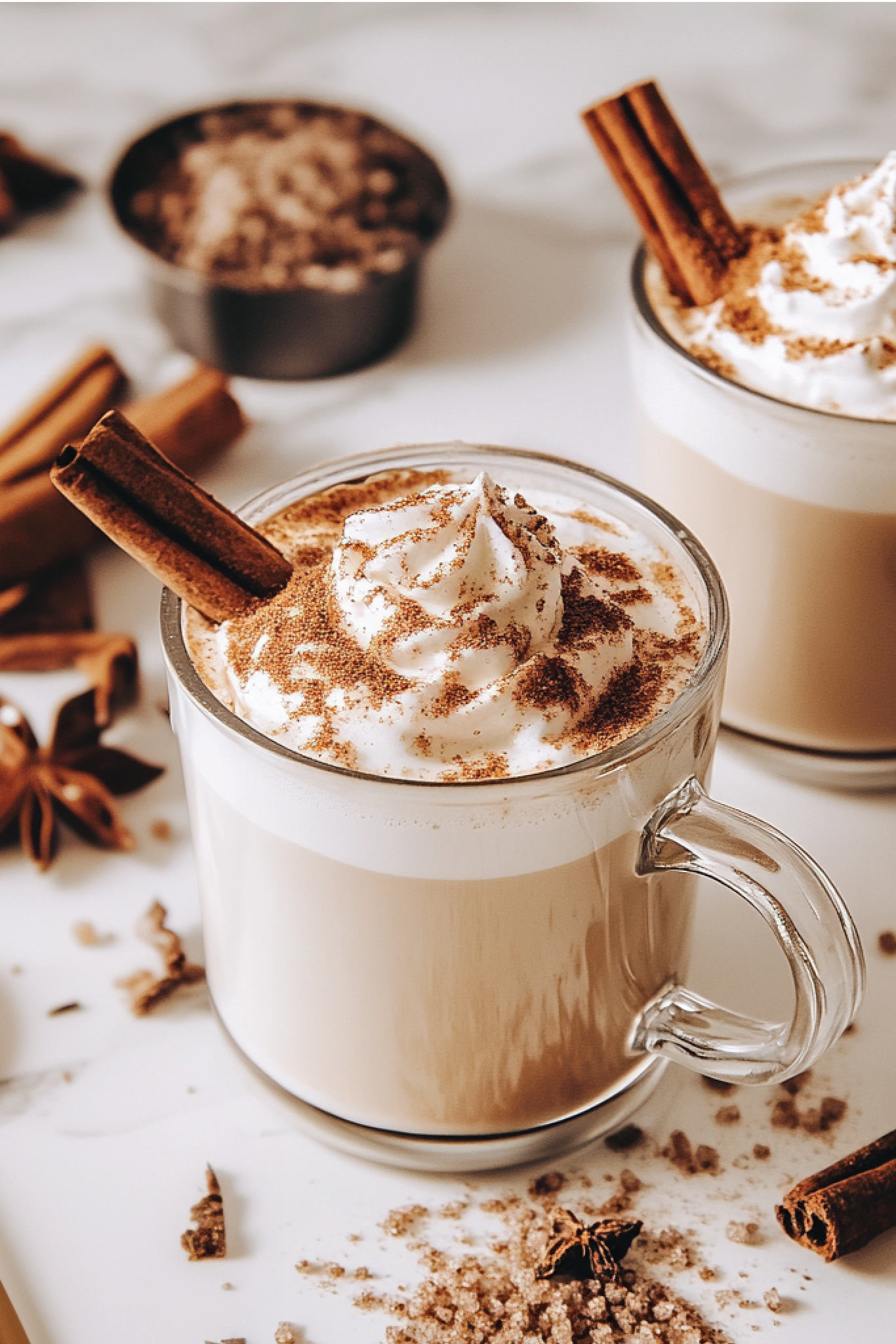 Two mugs of hot chocolate with whipped cream and cinnamon sticks on a marble surface.