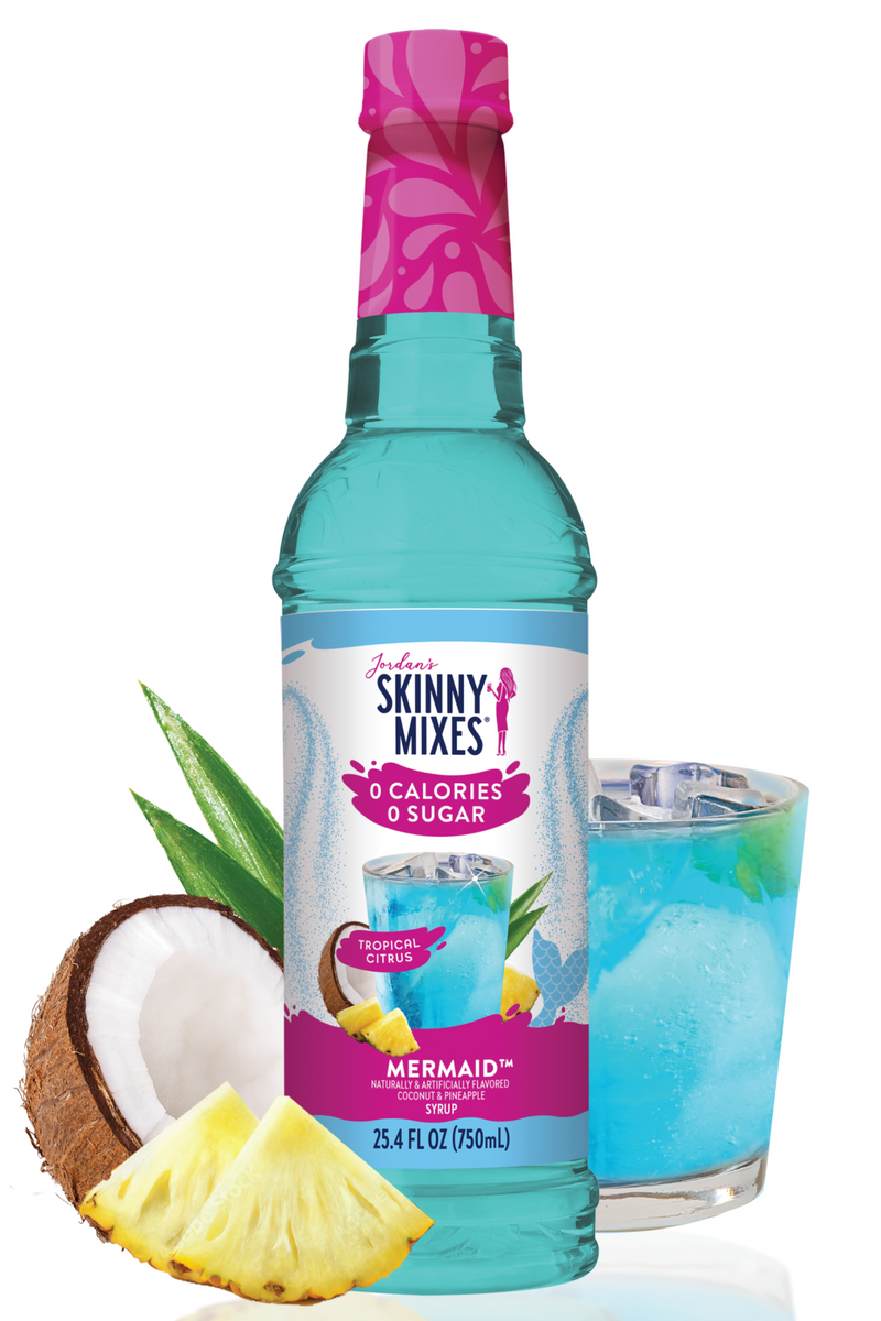 Bottle of Skinny Mixes Mermaid syrup with a glass of blue drink, pineapple, and coconut on a white background