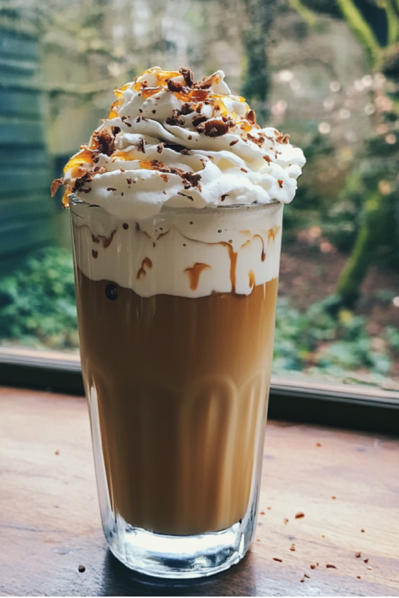 Cappuccino with whipped cream and chocolate shavings on a wooden table with a window in the background.