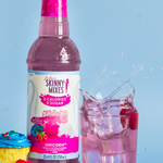 Bottle of Skinny Mixes Unicorn syrup with a glass of pink drink and cupcakes on a blue background