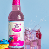 Bottle of Skinny Mixes Unicorn syrup with a glass of pink drink and cupcakes on a blue background