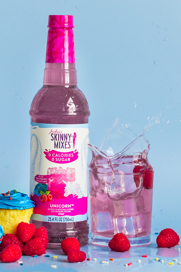 Bottle of Skinny Mixes Unicorn syrup with a glass of pink drink and cupcakes on a blue background