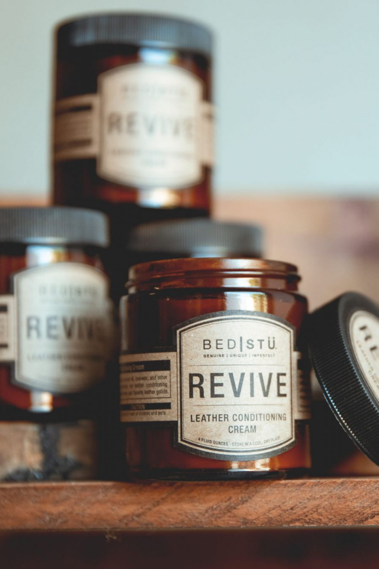 Multiple bottles of Revive Leather Conditioning Cream with brown labels on a wooden surface, with the primary bottle in the foreground.