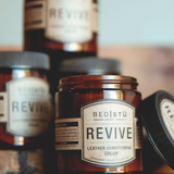 Multiple bottles of Revive Leather Conditioning Cream with brown labels on a wooden surface, with the primary bottle in the foreground.